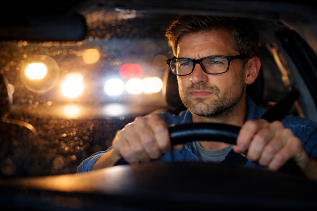 Man driving at night wearing glasses, squinting through glare and halos from oncoming headlights.