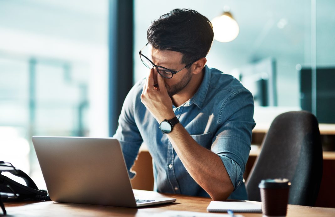 Man wearing glasses rubs the bridge of his nose while looking at a laptop, showing eye strain during screen time.