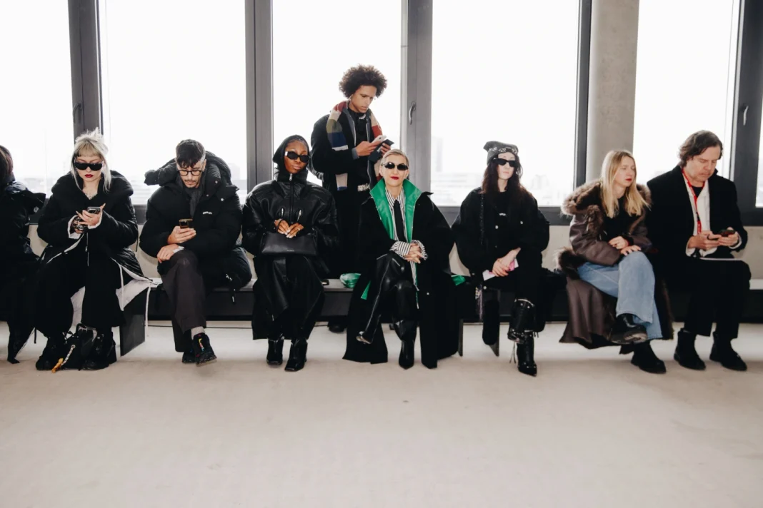 A front-row view of Berlin Fashion Week attendees sitting against a bright window, showcasing a mix of oversized black puffer coats, deconstructed leather jackets, and striking geometric eyewear. The aesthetic is cold and industrial, featuring a palette of stark blacks and a single pop of emerald green.