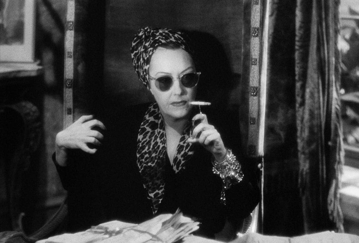 Norma Desmond wearing cat-eye sunglasses and a patterned headscarf while holding a cigarette in Sunset Boulevard.