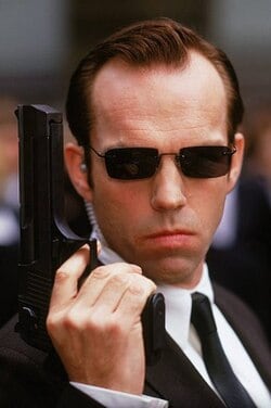 Agent Smith wearing black sunglasses and a black suit while holding a pistol in The Matrix.