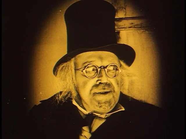 Sepia-toned still from the silent film The Cat and the Canary showing a man wearing a tall top hat and small round eyeglasses, lit dramatically against a dark background.