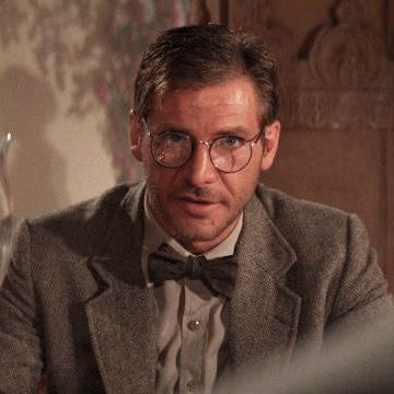 Close-up from an Indiana Jones film showing a man wearing small round eyeglasses, a gray tweed jacket, and a bow tie, looking intently toward someone off camera in a warmly lit room.