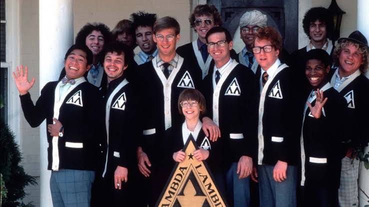 Group photo from Revenge of the Nerds featuring members of the Lambda Lambda Lambda fraternity wearing matching black-and-white cardigans, with several characters wearing eyeglasses, gathered around a fraternity sign outside a house.