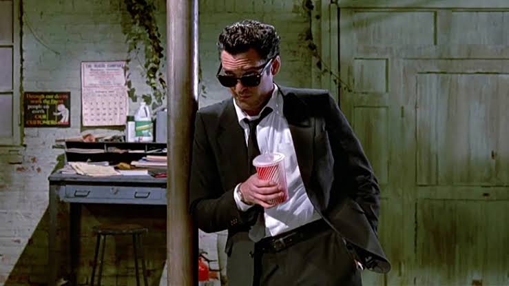Mr. Blonde wearing black sunglasses and a black suit while holding a drink in Reservoir Dogs.