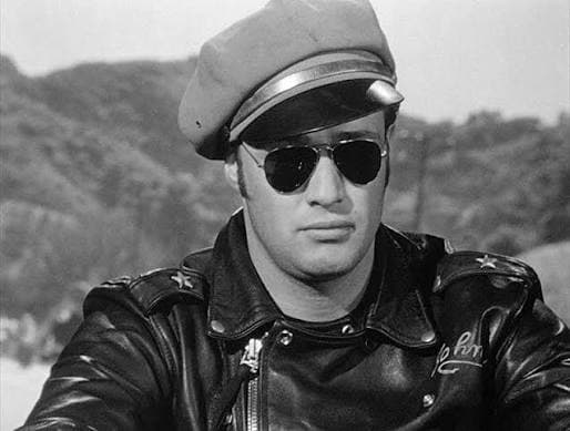 Johnny Strabler wearing aviator sunglasses, a leather jacket, and a cap while sitting on a motorcycle in The Wild One.