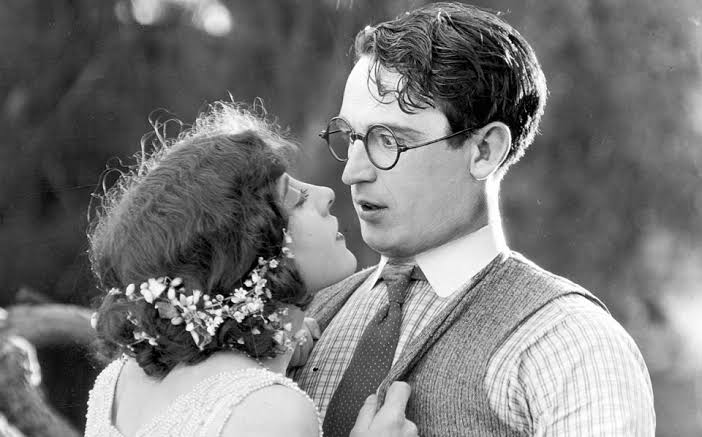 Black-and-white still from Girl Shy showing a young woman with flowers in her hair leaning close to a bespectacled young man with round eyeglasses. The man looks surprised as they gaze at each other outdoors.
