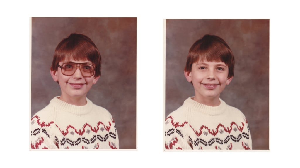 Child's school portrait shown in a side-by-side comparison, with large amber-tinted prescription glasses added to the left image and the original portrait without glasses on the right. The child wears a cream knit sweater with a red and black patterned design against a mottled brown studio background.